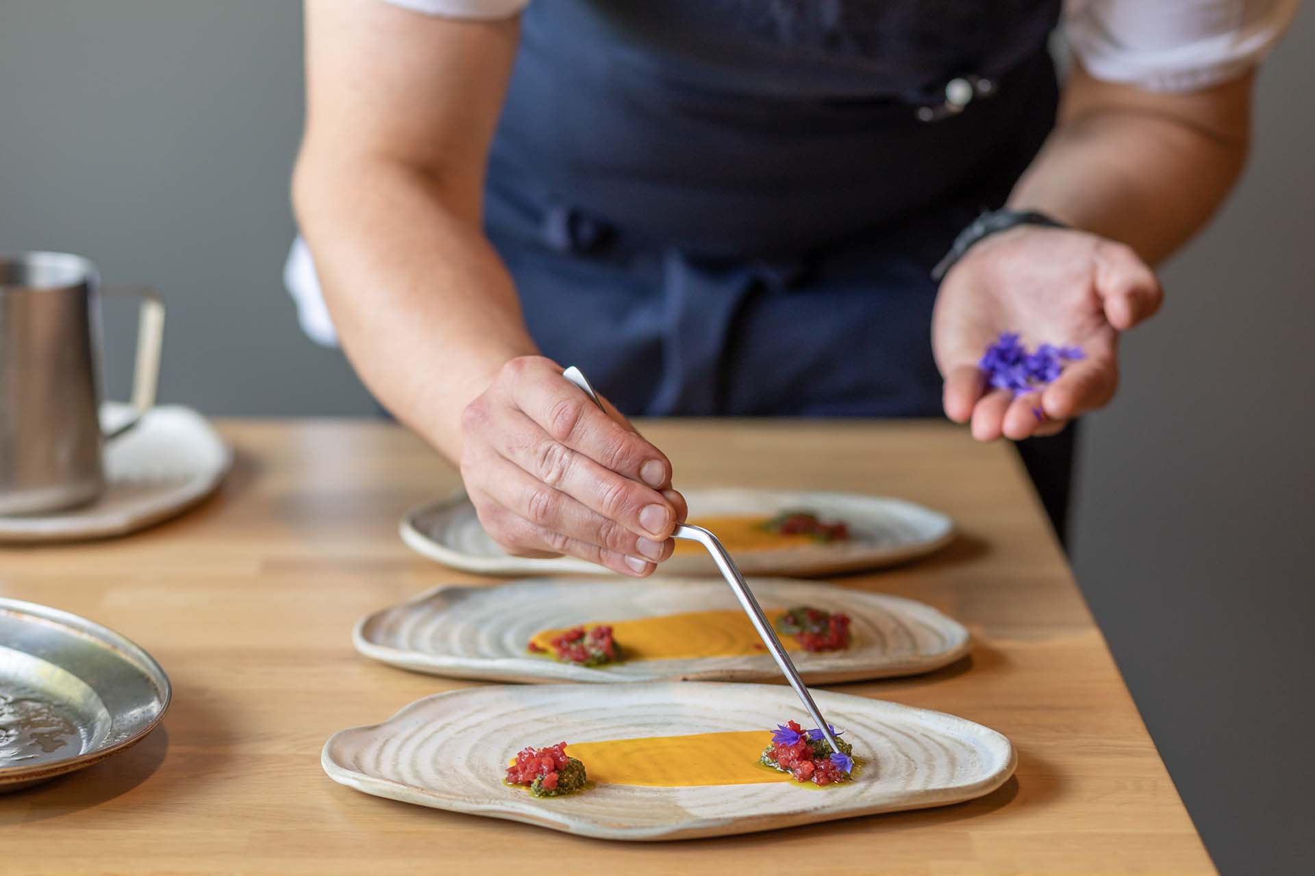 A chef stands at the end of a table. On the table are three plates in a line. Only the chef's hands and apron are visible, and the chef is depicted adding purple edible flowers to the plates with long tweezers. 